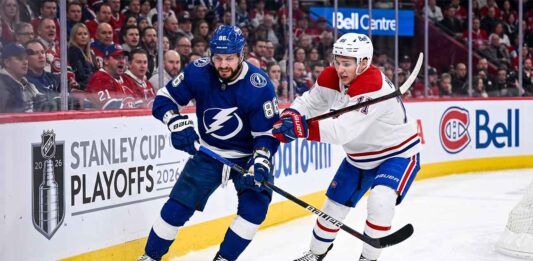 Dynamic on-ice action photo from Game 4 of the 2026 Stanley Cup Playoffs, showing Tampa Bay Lightning forward Nikita Kucherov (#86, blue jersey) battling Montreal Canadiens defenseman Lane Hutson (#?, white jersey) for puck possession near the boards in a packed Bell Centre.