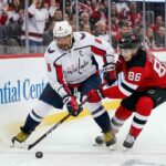 Washington Capitals captain Alex Ovechkin in an away white jersey battles New Jersey Devils star Jack Hughes for the puck during an NHL game.