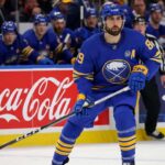 Buffalo Sabres forward Alex Tuch in action on the ice, skating past Coca-Cola and PS5 rink board advertisements during an NHL game.