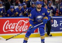 Buffalo Sabres forward Alex Tuch in action on the ice, skating past Coca-Cola and PS5 rink board advertisements during an NHL game.