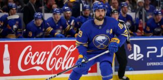 Buffalo Sabres forward Alex Tuch in action on the ice, skating past Coca-Cola and PS5 rink board advertisements during an NHL game.