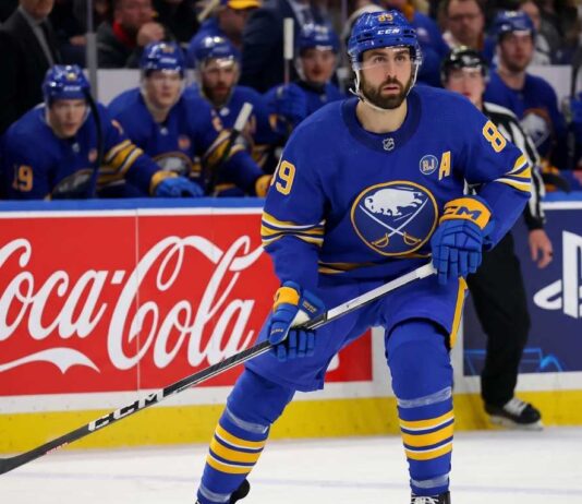 Buffalo Sabres forward Alex Tuch in action on the ice, skating past Coca-Cola and PS5 rink board advertisements during an NHL game.