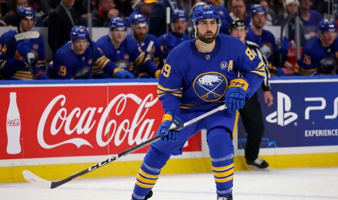 Buffalo Sabres forward Alex Tuch in action on the ice, skating past Coca-Cola and PS5 rink board advertisements during an NHL game.