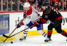A dynamic action photograph from the 2026 NHL Eastern Conference Finals Game 1, capturing Montreal Canadiens forward Cole Caufield fighting off Carolina Hurricanes forward Taylor Hall for possession of the hockey puck during intense, physical board play at Lenovo Center.