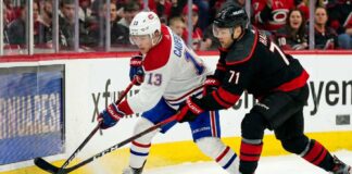 How to Watch Montreal Canadiens vs Carolina Hurricanes Game 2 on TV Today A dynamic action photograph from the 2026 NHL Eastern Conference Finals Game 1, capturing Montreal Canadiens forward Cole Caufield fighting off Carolina Hurricanes forward Taylor Hall for possession of the hockey puck during intense, physical board play at Lenovo Center.