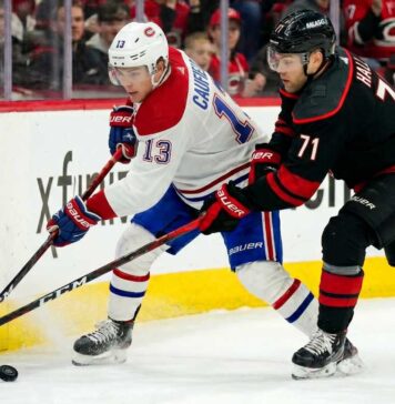 How to Watch Montreal Canadiens vs Carolina Hurricanes Game 2 on TV Today A dynamic action photograph from the 2026 NHL Eastern Conference Finals Game 1, capturing Montreal Canadiens forward Cole Caufield fighting off Carolina Hurricanes forward Taylor Hall for possession of the hockey puck during intense, physical board play at Lenovo Center.