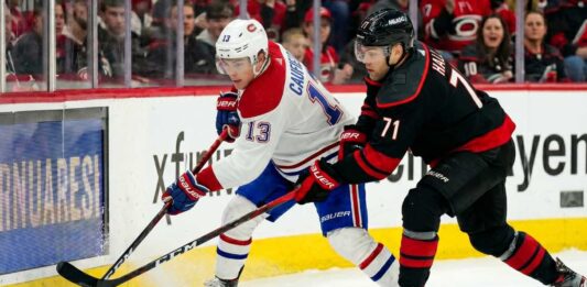 A dynamic action photograph from the 2026 NHL Eastern Conference Finals Game 1, capturing Montreal Canadiens forward Cole Caufield fighting off Carolina Hurricanes forward Taylor Hall for possession of the hockey puck during intense, physical board play at Lenovo Center.