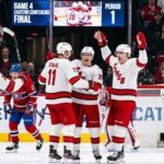 Carolina Hurricanes and Montreal Canadiens Game 4 Recap & Score A group of Carolina Hurricanes players in white away jerseys, including Sebastian Aho (20) and Jordan Staal (11), huddle on the ice in triumph, celebrating a first-period goal against the Montreal Canadiens during Game 4 of the Eastern Conference Final. In the background, a large Jumbotron confirms the event and period.