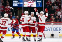 A group of Carolina Hurricanes players in white away jerseys, including Sebastian Aho (20) and Jordan Staal (11), huddle on the ice in triumph, celebrating a first-period goal against the Montreal Canadiens during Game 4 of the Eastern Conference Final. In the background, a large Jumbotron confirms the event and period.