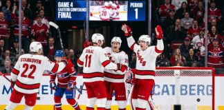 A group of Carolina Hurricanes players in white away jerseys, including Sebastian Aho (20) and Jordan Staal (11), huddle on the ice in triumph, celebrating a first-period goal against the Montreal Canadiens during Game 4 of the Eastern Conference Final. In the background, a large Jumbotron confirms the event and period.