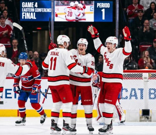 Carolina Hurricanes and Montreal Canadiens Game 4 Recap & Score A group of Carolina Hurricanes players in white away jerseys, including Sebastian Aho (20) and Jordan Staal (11), huddle on the ice in triumph, celebrating a first-period goal against the Montreal Canadiens during Game 4 of the Eastern Conference Final. In the background, a large Jumbotron confirms the event and period.