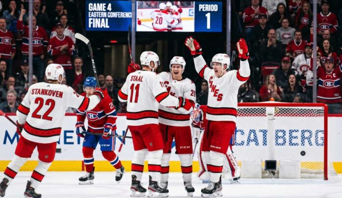 carolina-hurricanes-montreal-canadiens-game-4-shutout A group of Carolina Hurricanes players in white away jerseys, including Sebastian Aho (20) and Jordan Staal (11), huddle on the ice in triumph, celebrating a first-period goal against the Montreal Canadiens during Game 4 of the Eastern Conference Final. In the background, a large Jumbotron confirms the event and period.
