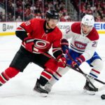 Carolina Hurricanes’ Sebastian Aho (jersey 20) and Montreal Canadiens’ Lane Hutson (jersey 21) battle intensely for a hockey puck on the ice during Game 4 of the 2026 Eastern Conference Final. Both players are low to the ice, locking sticks over the puck, with a crowded arena and hockey boards visible in the background.