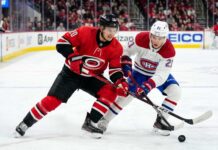How to Watch Carolina Hurricanes vs Montreal Canadiens Game 4 Today Carolina Hurricanes’ Sebastian Aho (jersey 20) and Montreal Canadiens’ Lane Hutson (jersey 21) battle intensely for a hockey puck on the ice during Game 4 of the 2026 Eastern Conference Final. Both players are low to the ice, locking sticks over the puck, with a crowded arena and hockey boards visible in the background.