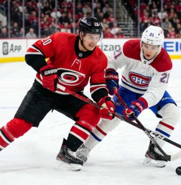 Carolina Hurricanes’ Sebastian Aho (jersey 20) and Montreal Canadiens’ Lane Hutson (jersey 21) battle intensely for a hockey puck on the ice during Game 4 of the 2026 Eastern Conference Final. Both players are low to the ice, locking sticks over the puck, with a crowded arena and hockey boards visible in the background.