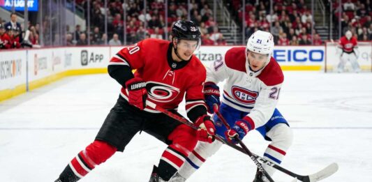 Carolina Hurricanes’ Sebastian Aho (jersey 20) and Montreal Canadiens’ Lane Hutson (jersey 21) battle intensely for a hockey puck on the ice during Game 4 of the 2026 Eastern Conference Final. Both players are low to the ice, locking sticks over the puck, with a crowded arena and hockey boards visible in the background.
