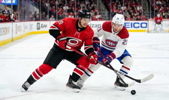 carolina-hurricanes-montreal-canadiens-playoffs-game-4 Carolina Hurricanes’ Sebastian Aho (jersey 20) and Montreal Canadiens’ Lane Hutson (jersey 21) battle intensely for a hockey puck on the ice during Game 4 of the 2026 Eastern Conference Final. Both players are low to the ice, locking sticks over the puck, with a crowded arena and hockey boards visible in the background.
