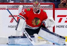 A photorealistic image showing goalie Connor Hellebuyck in a red Chicago Blackhawks home uniform and custom Blackhawks mask in the goal crease, illustrating potential trade rumors from the Winnipeg Jets.