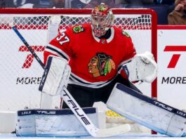 A photorealistic image showing goalie Connor Hellebuyck in a red Chicago Blackhawks home uniform and custom Blackhawks mask in the goal crease, illustrating potential trade rumors from the Winnipeg Jets.