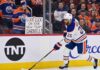 Edmonton Oilers defenseman Darnell Nurse skating on the ice with a fan sign in the background reading 'GOOD LUCK ON YOUR NEW TEAM DARNELL!' during intense offseason trade rumors.