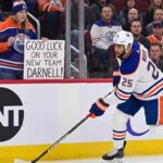 Edmonton Oilers defenseman Darnell Nurse skating on the ice with a fan sign in the background reading 'GOOD LUCK ON YOUR NEW TEAM DARNELL!' during intense offseason trade rumors.