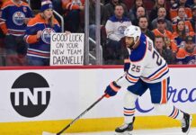 Edmonton Oilers defenseman Darnell Nurse skating on the ice with a fan sign in the background reading 'GOOD LUCK ON YOUR NEW TEAM DARNELL!' during intense offseason trade rumors.