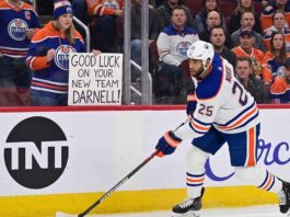 Edmonton Oilers defenseman Darnell Nurse skating on the ice with a fan sign in the background reading 'GOOD LUCK ON YOUR NEW TEAM DARNELL!' during intense offseason trade rumors.