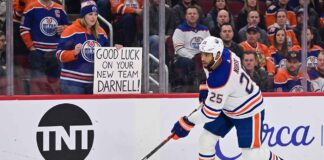 Edmonton Oilers defenseman Darnell Nurse skating on the ice with a fan sign in the background reading 'GOOD LUCK ON YOUR NEW TEAM DARNELL!' during intense offseason trade rumors.
