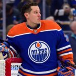 Photo-realistic mockup of Jordan Binnington wearing an Edmonton Oilers uniform and holding an Oilers-themed goalie mask, illustrating NHL trade rumors between the St. Louis Blues and Edmonton.