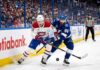 A dynamic photograph showing Montreal Canadiens defenseman Lane Hutson (left, #20) and Tampa Bay Lightning winger Nikita Kucherov (right, #86) locked in an intense battle for the puck on the ice during Game 7 of the 2026 Stanley Cup Playoffs. They are entangled with sticks low, generating ice spray, against a blurred background of the arena crowd and Scotiabank boards.