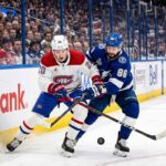 A dynamic photograph showing Montreal Canadiens defenseman Lane Hutson (left, #20) and Tampa Bay Lightning winger Nikita Kucherov (right, #86) locked in an intense battle for the puck on the ice during Game 7 of the 2026 Stanley Cup Playoffs. They are entangled with sticks low, generating ice spray, against a blurred background of the arena crowd and Scotiabank boards.