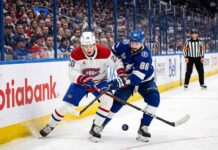 A dynamic photograph showing Montreal Canadiens defenseman Lane Hutson (left, #20) and Tampa Bay Lightning winger Nikita Kucherov (right, #86) locked in an intense battle for the puck on the ice during Game 7 of the 2026 Stanley Cup Playoffs. They are entangled with sticks low, generating ice spray, against a blurred background of the arena crowd and Scotiabank boards.