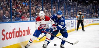 A dynamic photograph showing Montreal Canadiens defenseman Lane Hutson (left, #20) and Tampa Bay Lightning winger Nikita Kucherov (right, #86) locked in an intense battle for the puck on the ice during Game 7 of the 2026 Stanley Cup Playoffs. They are entangled with sticks low, generating ice spray, against a blurred background of the arena crowd and Scotiabank boards.