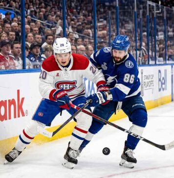 A dynamic photograph showing Montreal Canadiens defenseman Lane Hutson (left, #20) and Tampa Bay Lightning winger Nikita Kucherov (right, #86) locked in an intense battle for the puck on the ice during Game 7 of the 2026 Stanley Cup Playoffs. They are entangled with sticks low, generating ice spray, against a blurred background of the arena crowd and Scotiabank boards.