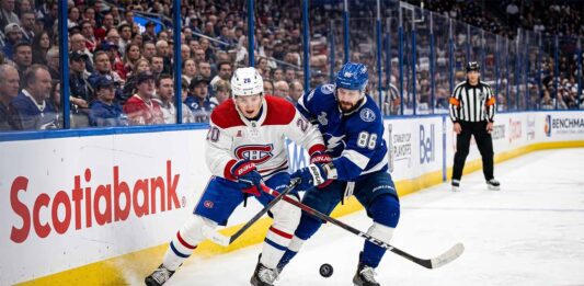 A dynamic photograph showing Montreal Canadiens defenseman Lane Hutson (left, #20) and Tampa Bay Lightning winger Nikita Kucherov (right, #86) locked in an intense battle for the puck on the ice during Game 7 of the 2026 Stanley Cup Playoffs. They are entangled with sticks low, generating ice spray, against a blurred background of the arena crowd and Scotiabank boards.