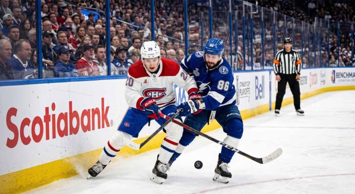 A dynamic photograph showing Montreal Canadiens defenseman Lane Hutson (left, #20) and Tampa Bay Lightning winger Nikita Kucherov (right, #86) locked in an intense battle for the puck on the ice during Game 7 of the 2026 Stanley Cup Playoffs. They are entangled with sticks low, generating ice spray, against a blurred background of the arena crowd and Scotiabank boards.