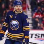 Jake DeBrusk Trade Rumors: Will the Canucks Deal Him to the Sabres? A detailed photo-realistic visualization of forward Jake DeBrusk, featuring him in a dark navy and gold Buffalo Sabres uniform with the crossed-swords-and-buffalo logo. His specific number 74 is clearly visible on his helmet, jersey sleeves, and chest. He holds a Bauer hockey stick on the ice during an NHL game visualization, set against a blurred arena background with visible advertising.