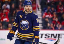 A detailed photo-realistic visualization of forward Jake DeBrusk, featuring him in a dark navy and gold Buffalo Sabres uniform with the crossed-swords-and-buffalo logo. His specific number 74 is clearly visible on his helmet, jersey sleeves, and chest. He holds a Bauer hockey stick on the ice during an NHL game visualization, set against a blurred arena background with visible advertising.