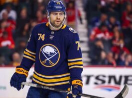 Jake DeBrusk Trade Rumors: Will the Canucks Deal Him to the Sabres? A detailed photo-realistic visualization of forward Jake DeBrusk, featuring him in a dark navy and gold Buffalo Sabres uniform with the crossed-swords-and-buffalo logo. His specific number 74 is clearly visible on his helmet, jersey sleeves, and chest. He holds a Bauer hockey stick on the ice during an NHL game visualization, set against a blurred arena background with visible advertising.