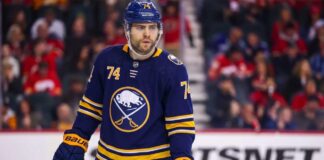 A detailed photo-realistic visualization of forward Jake DeBrusk, featuring him in a dark navy and gold Buffalo Sabres uniform with the crossed-swords-and-buffalo logo. His specific number 74 is clearly visible on his helmet, jersey sleeves, and chest. He holds a Bauer hockey stick on the ice during an NHL game visualization, set against a blurred arena background with visible advertising.