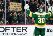 A photo of Minnesota Wild goaltender Jesper Wallstedt (30) in his green and gold alternate jersey celebrating on the ice as a fan in the stands holds a sign that reads 'Good luck on your new team Jesper!' during an NHL game, highlighting potential trade rumors.