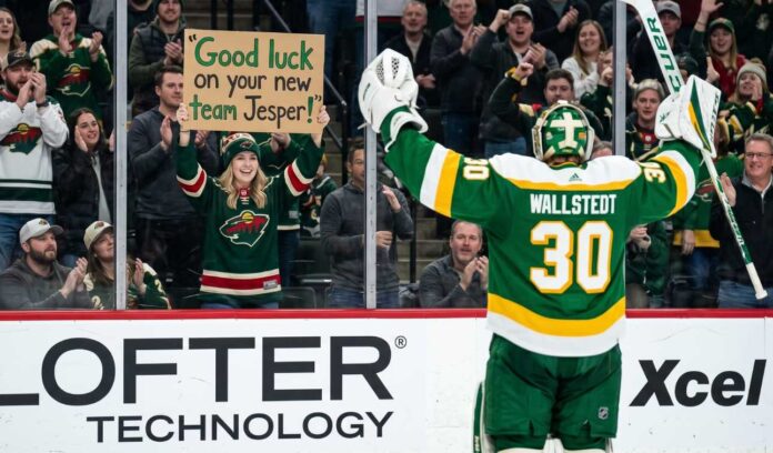 A photo of Minnesota Wild goaltender Jesper Wallstedt (30) in his green and gold alternate jersey celebrating on the ice as a fan in the stands holds a sign that reads 'Good luck on your new team Jesper!' during an NHL game, highlighting potential trade rumors.