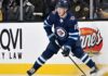 A close-up action shot on ice of hockey player Pavel Dorofeyev, featuring his likeness and low-crouched pose, now in a navy blue Winnipeg Jets home uniform and helmet with number 16, controlling a puck with his stick and creating ice spray.