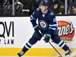 A close-up action shot on ice of hockey player Pavel Dorofeyev, featuring his likeness and low-crouched pose, now in a navy blue Winnipeg Jets home uniform and helmet with number 16, controlling a puck with his stick and creating ice spray.