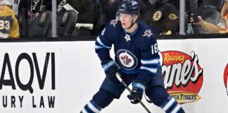 A close-up action shot on ice of hockey player Pavel Dorofeyev, featuring his likeness and low-crouched pose, now in a navy blue Winnipeg Jets home uniform and helmet with number 16, controlling a puck with his stick and creating ice spray.