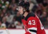 Close-up profile photograph of NHL star Quinn Hughes on the ice, wearing a New Jersey Devils home uniform and black helmet to visualize trade rumors that would reunite him with his brothers.