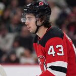 Quinn Hughes Trade Rumors: Minnesota Wild to New Jersey Devils? Close-up profile photograph of NHL star Quinn Hughes on the ice, wearing a New Jersey Devils home uniform and black helmet to visualize trade rumors that would reunite him with his brothers.