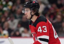 Quinn Hughes Trade Rumors: Minnesota Wild to New Jersey Devils? Close-up profile photograph of NHL star Quinn Hughes on the ice, wearing a New Jersey Devils home uniform and black helmet to visualize trade rumors that would reunite him with his brothers.