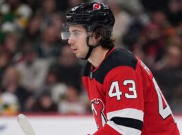 Quinn Hughes Trade Rumors: Minnesota Wild to New Jersey Devils? Close-up profile photograph of NHL star Quinn Hughes on the ice, wearing a New Jersey Devils home uniform and black helmet to visualize trade rumors that would reunite him with his brothers.