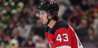 Quinn Hughes Trade Rumors: Minnesota Wild to New Jersey Devils? Close-up profile photograph of NHL star Quinn Hughes on the ice, wearing a New Jersey Devils home uniform and black helmet to visualize trade rumors that would reunite him with his brothers.